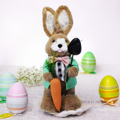 Coelho de pelúcia decorativo vestido com roupa social verde, segurando uma cenoura e uma enxada, ideal para decoração de Páscoa e ambientes rústicos.