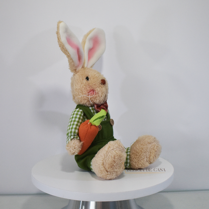 Coelho de pelúcia bege, vestido com macacão verde e camisa xadrez, segurando uma cenoura, ideal para decoração de Páscoa e ambientes temáticos.