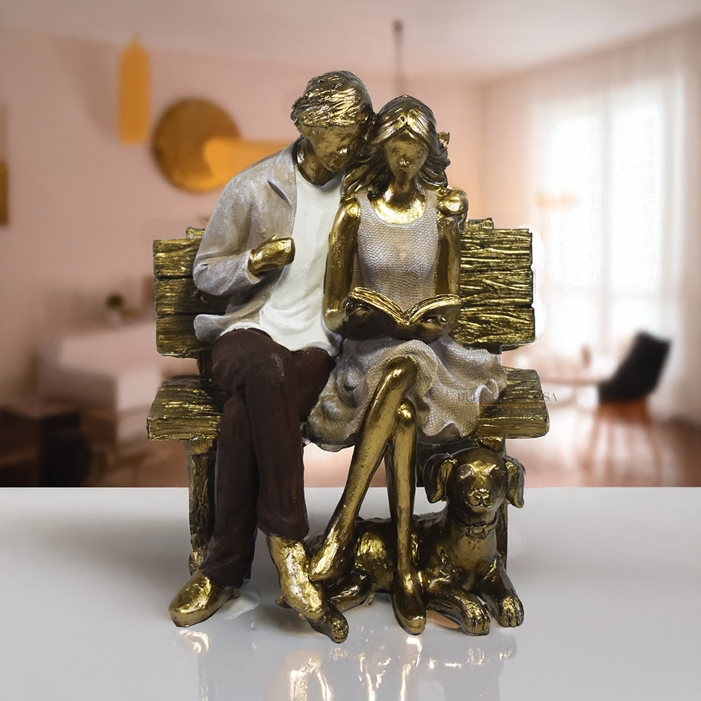 Escultura decorativa em resina de casal sentado em banco lendo um livro, acompanhados por um cachorro aos pés, com acabamento dourado, branco e marrom. Peça elegante para decoração de interiores medindo 15x10x18 cm.