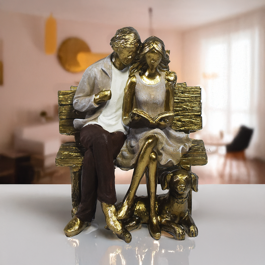 Escultura decorativa em resina de casal sentado em banco lendo um livro, acompanhados por um cachorro aos pés, com acabamento dourado, branco e marrom. Peça elegante para decoração de interiores medindo 15x10x18 cm.