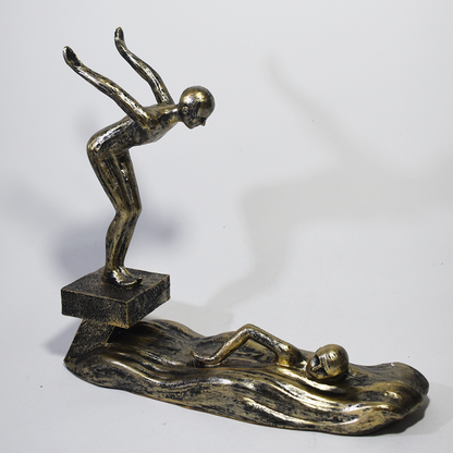 Escultura decorativa em resina representando atletas da natação, com um mergulhador e um nadador sobre ondas estilizadas em acabamento dourado envelhecido.