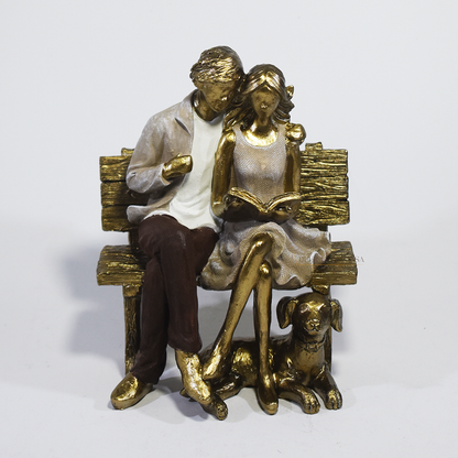 Escultura decorativa em resina de casal sentado em banco lendo um livro, acompanhados por um cachorro aos pés, com acabamento dourado, branco e marrom. Peça elegante para decoração de interiores medindo 15x10x18 cm.