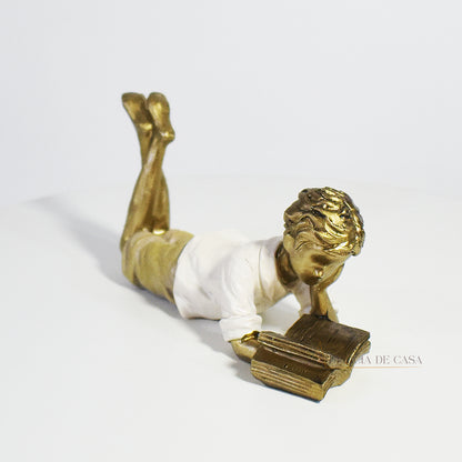 Escultura decorativa de menino lendo, deitada com as pernas cruzadas, feita em resina dourada com vestido em tons claros. Decoração para apaixonados por livros.

