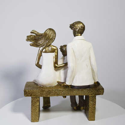Escultura decorativa em resina representando pai, mãe e filho sentados em um banco lendo um livro, simbolizando união familiar e momentos de aprendizado.