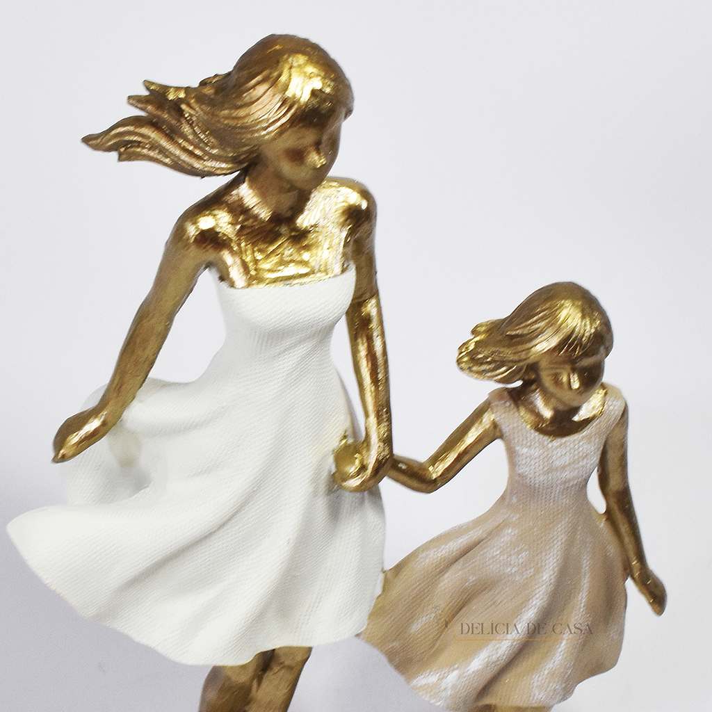 Estatueta decorativa em resina representando mãe e filha de mãos dadas, com acabamento dourado e vestidos em tons suaves. Peça elegante para simbolizar amor e conexão familiar na decoração.