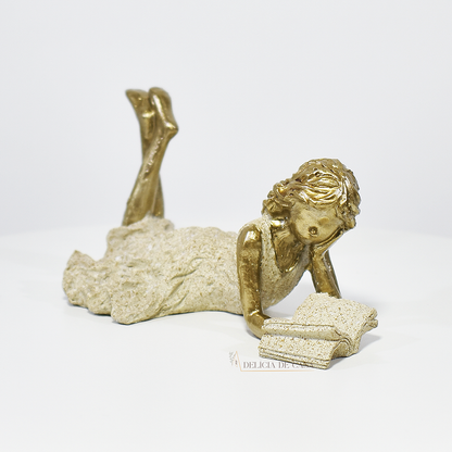 Escultura decorativa de menina lendo, deitada com as pernas cruzadas, feita em resina dourada com vestido em tons claros. Decoração para apaixonados por livros.